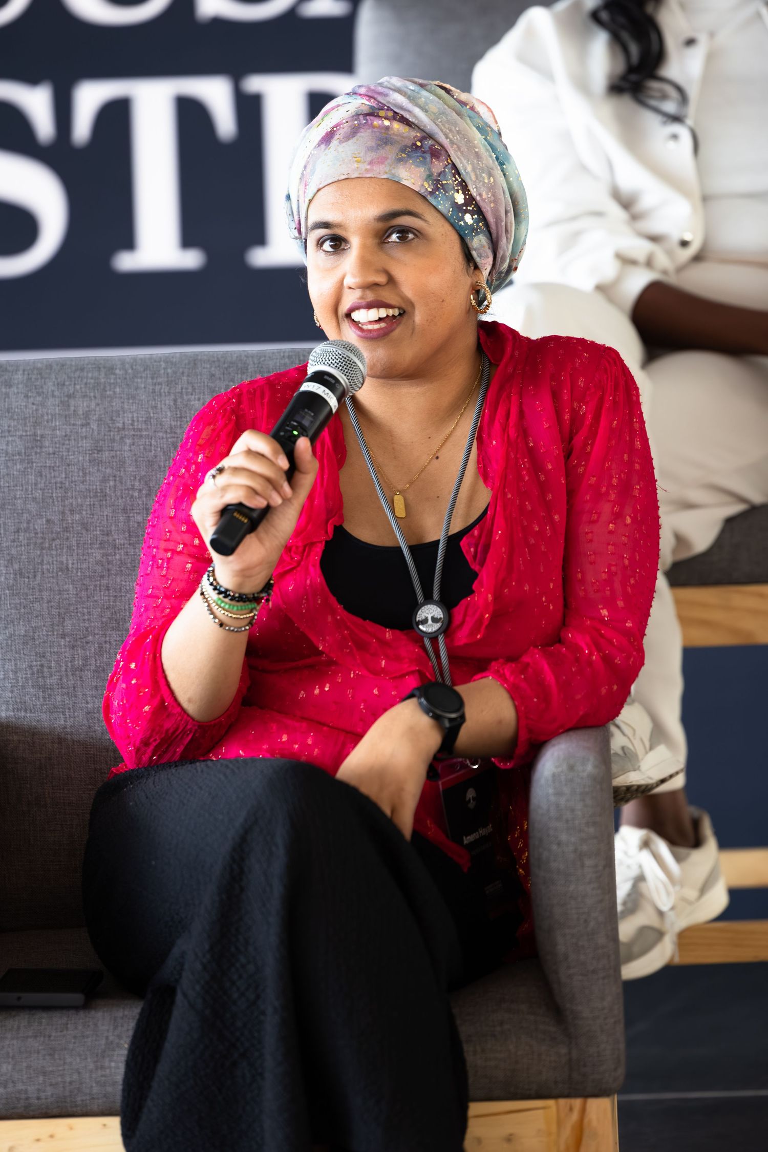 Speaker in a bright red top addresses audience while seated on stage during presentation.