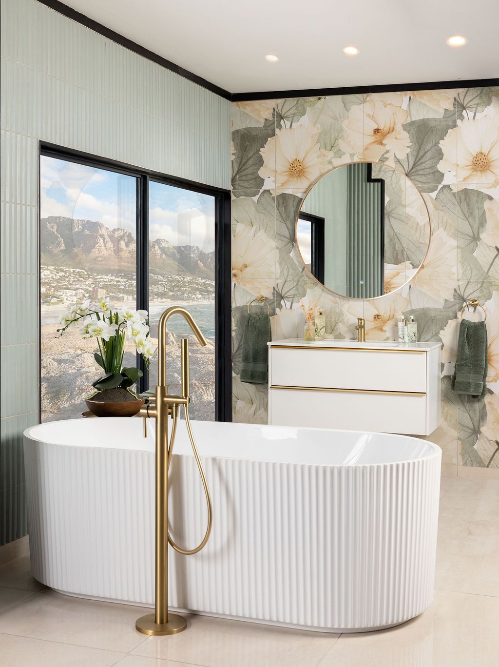 Luxurious modern bathroom featuring a freestanding white ribbed tub with gold fixtures and floral wallpaper against mountain views.
