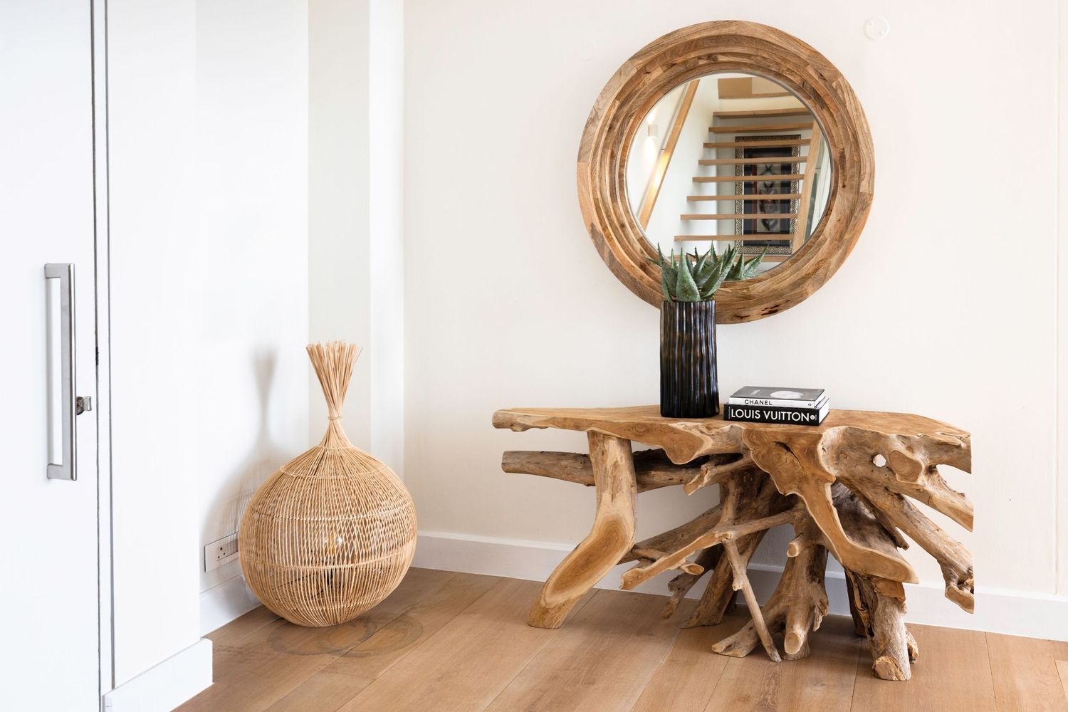 Rustic wooden console table with round mirror and natural woven decor elements in white hallway.