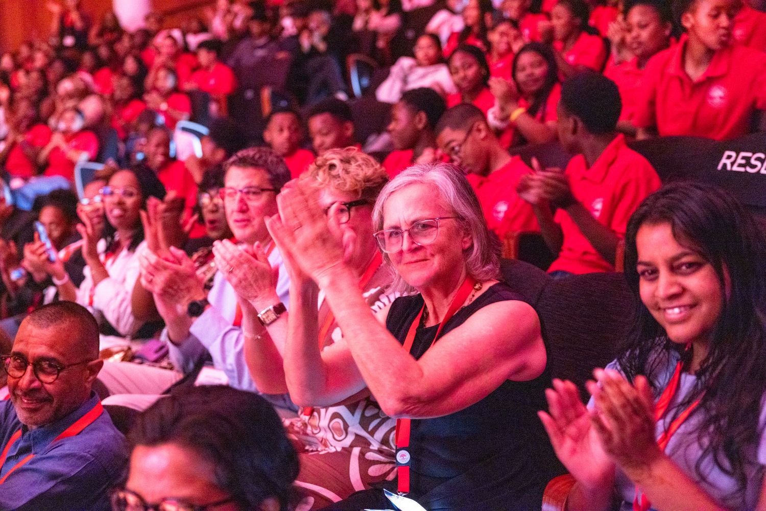 Audience members in red lighting watch intently during an indoor event or performance.