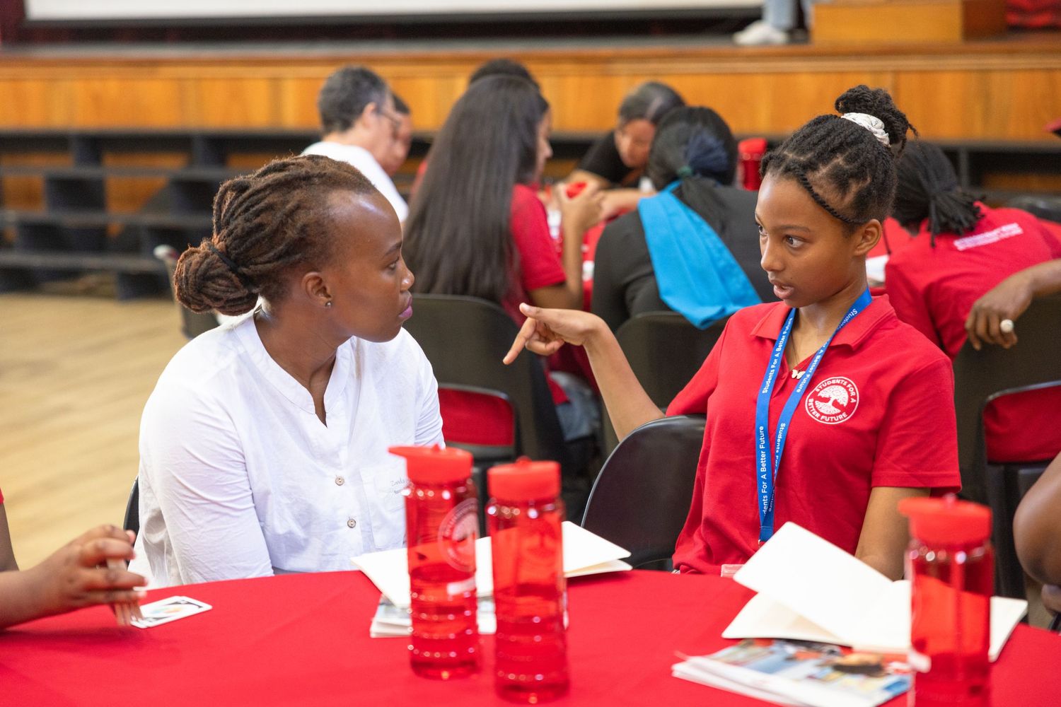 Two participants have a focused conversation at a table with red tablecloths and water bottles.