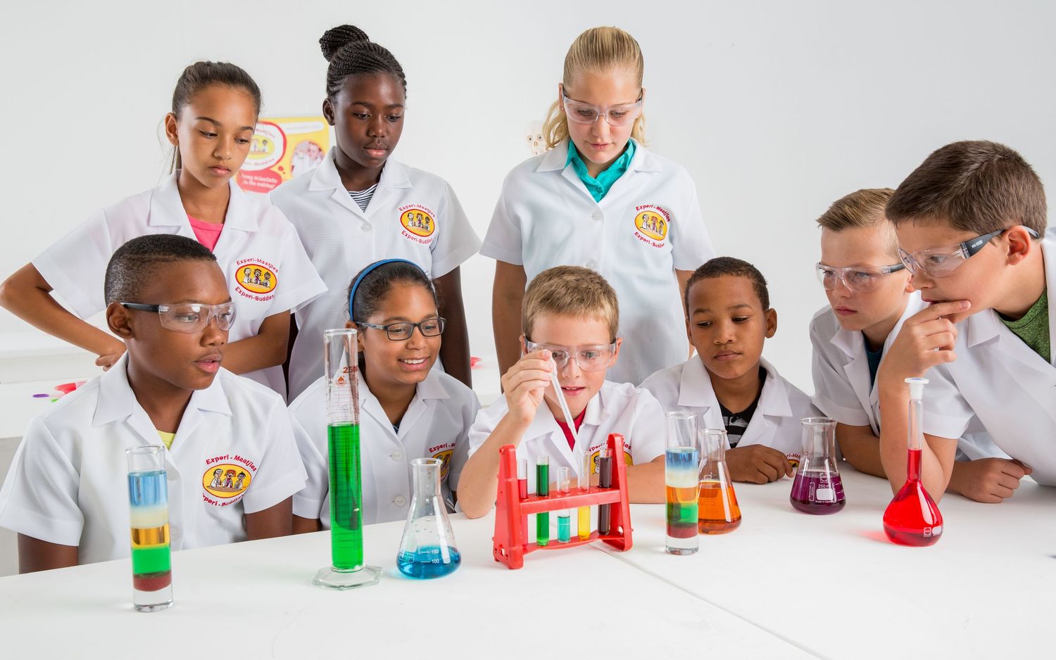 Group of students in lab coats observe colorful chemistry experiments on white table.