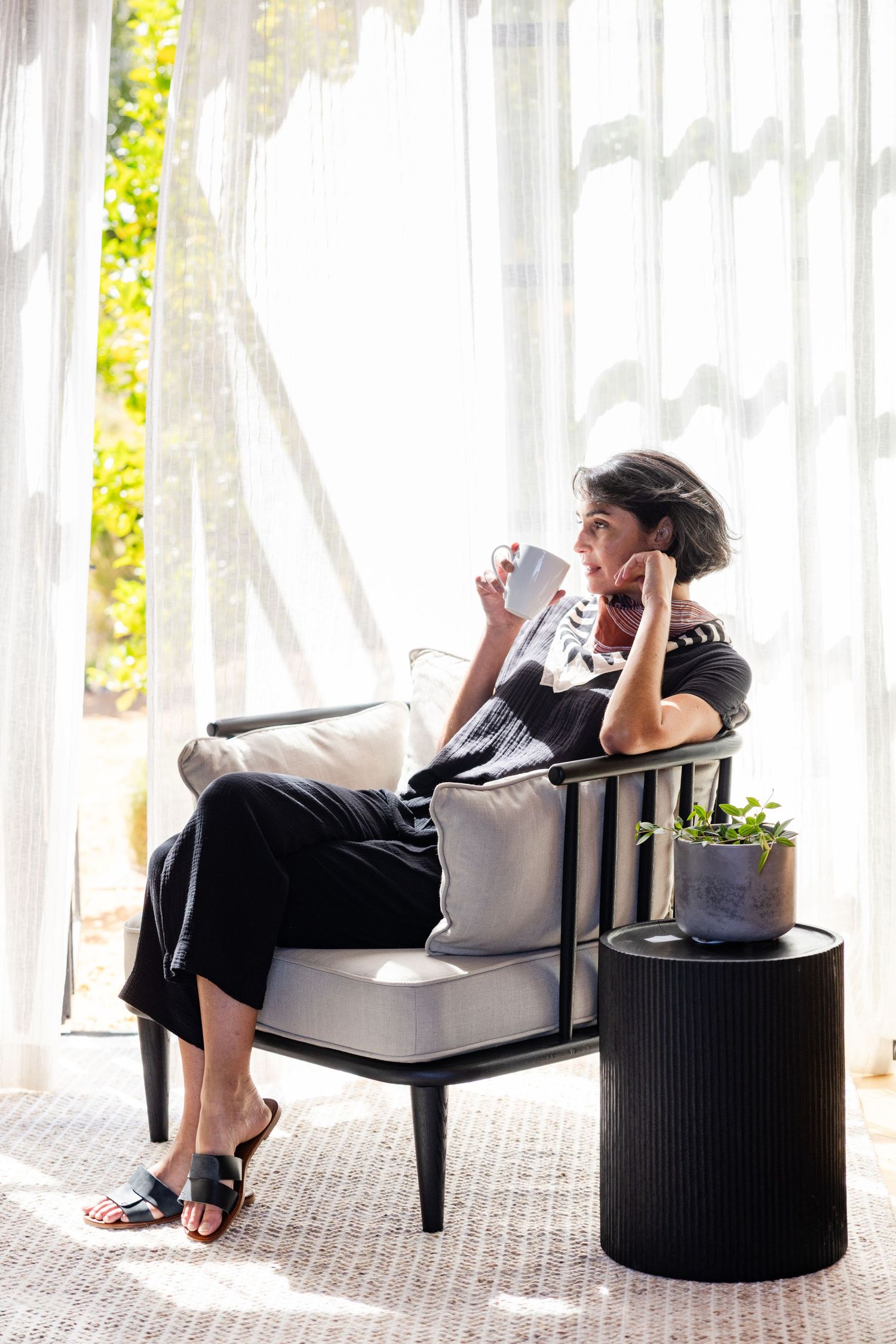 Person relaxing on modern patio chair by sheer white curtains and decorative plant.