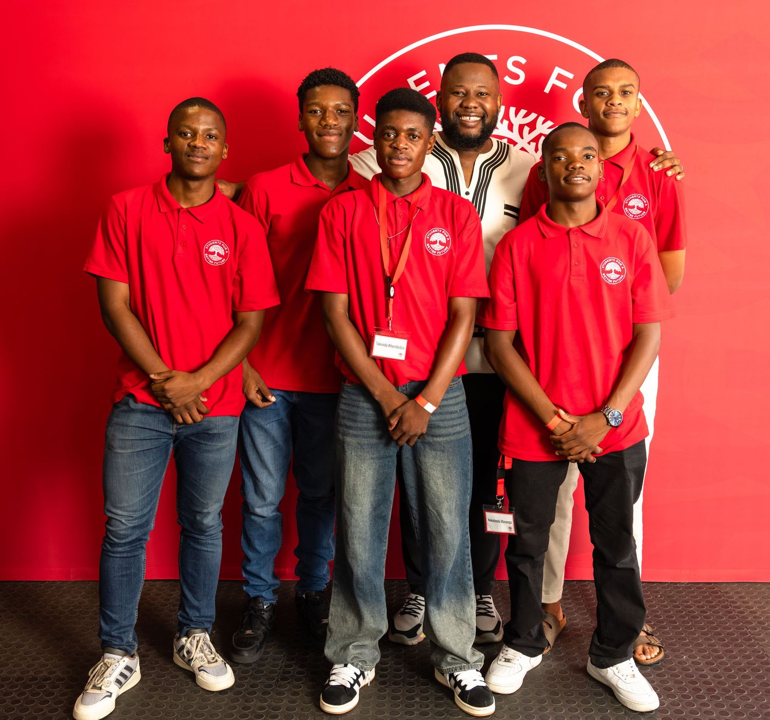 Group of students wearing matching red shirts pose together against red background with logo.