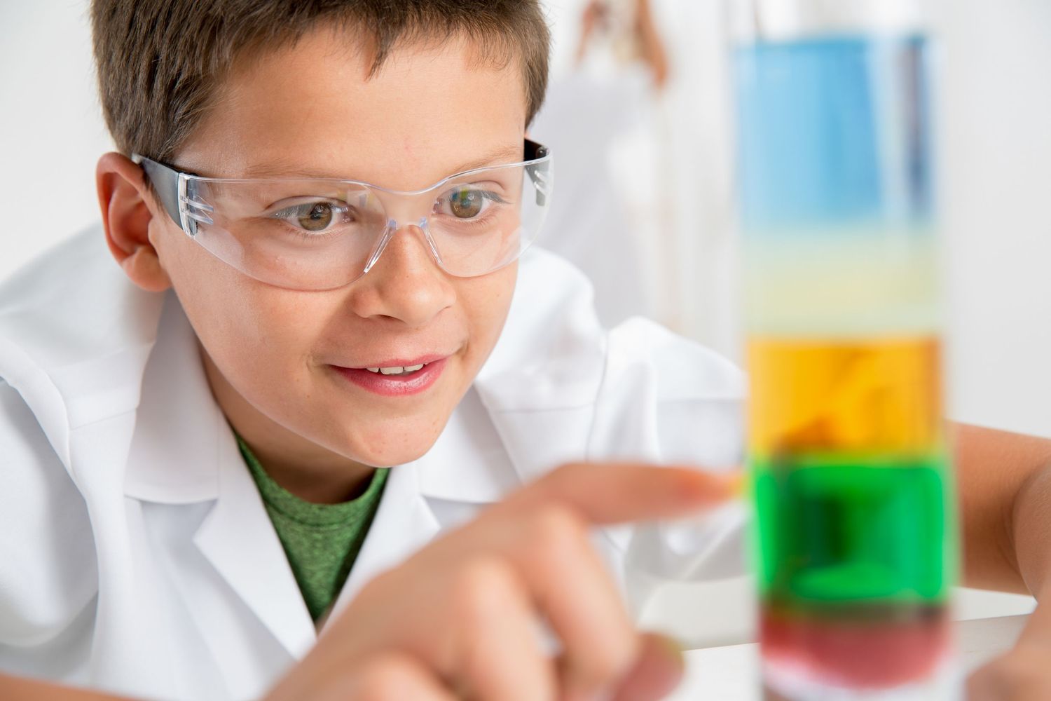 Student wearing safety glasses observes colorful layered liquids in science experiment.