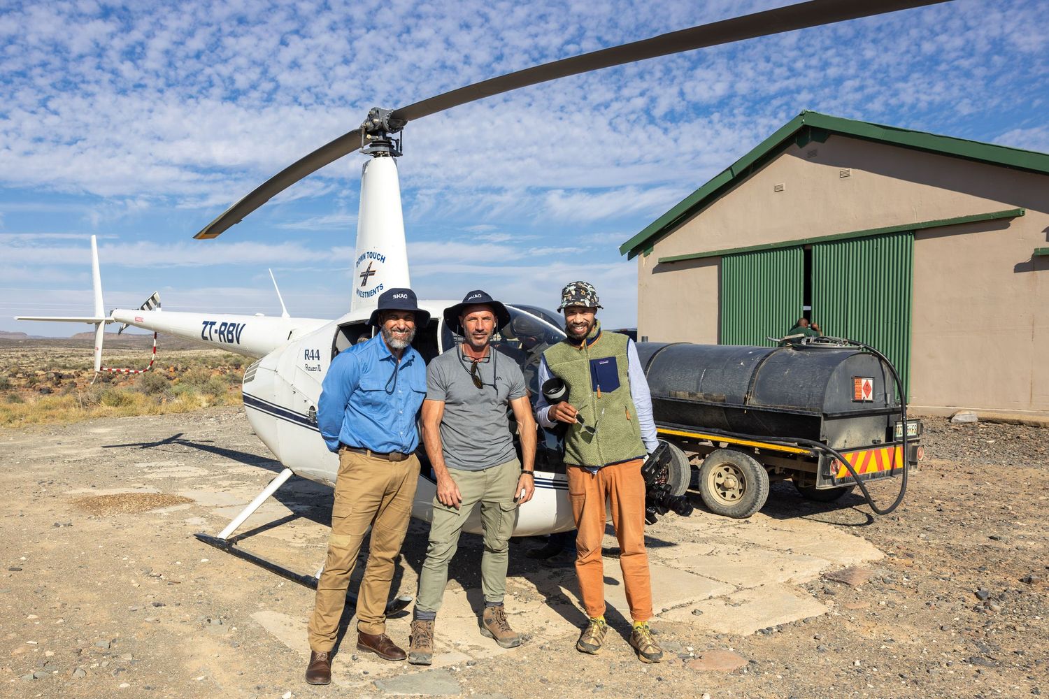 Three people standing in front of helicopter on desert landing pad.