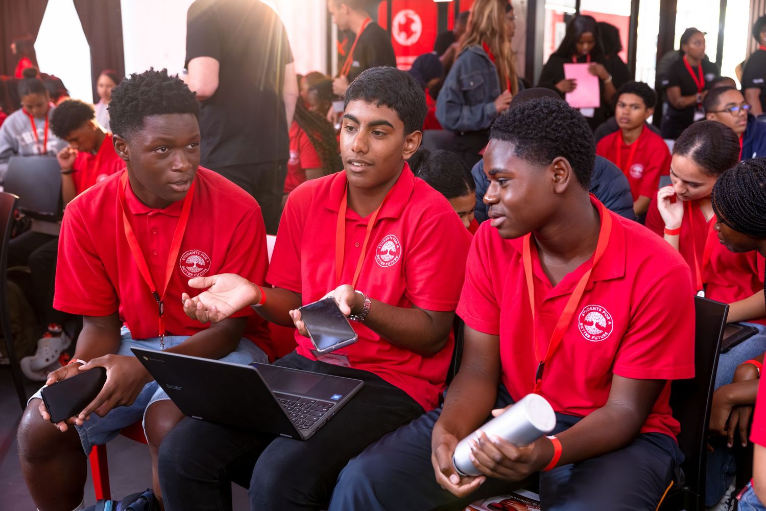 Three students in red polo shirts engage in discussion during a conference session.