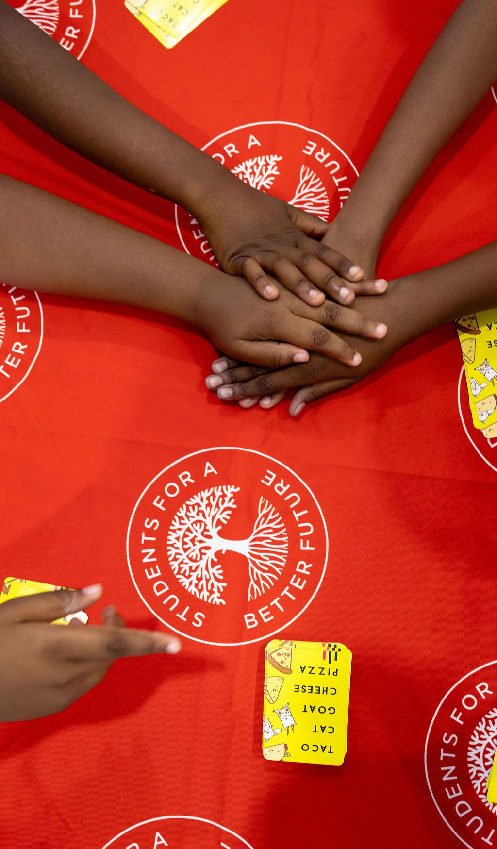 Hands placed together on red background with circular logo and yellow cards showing unity and collaboration.