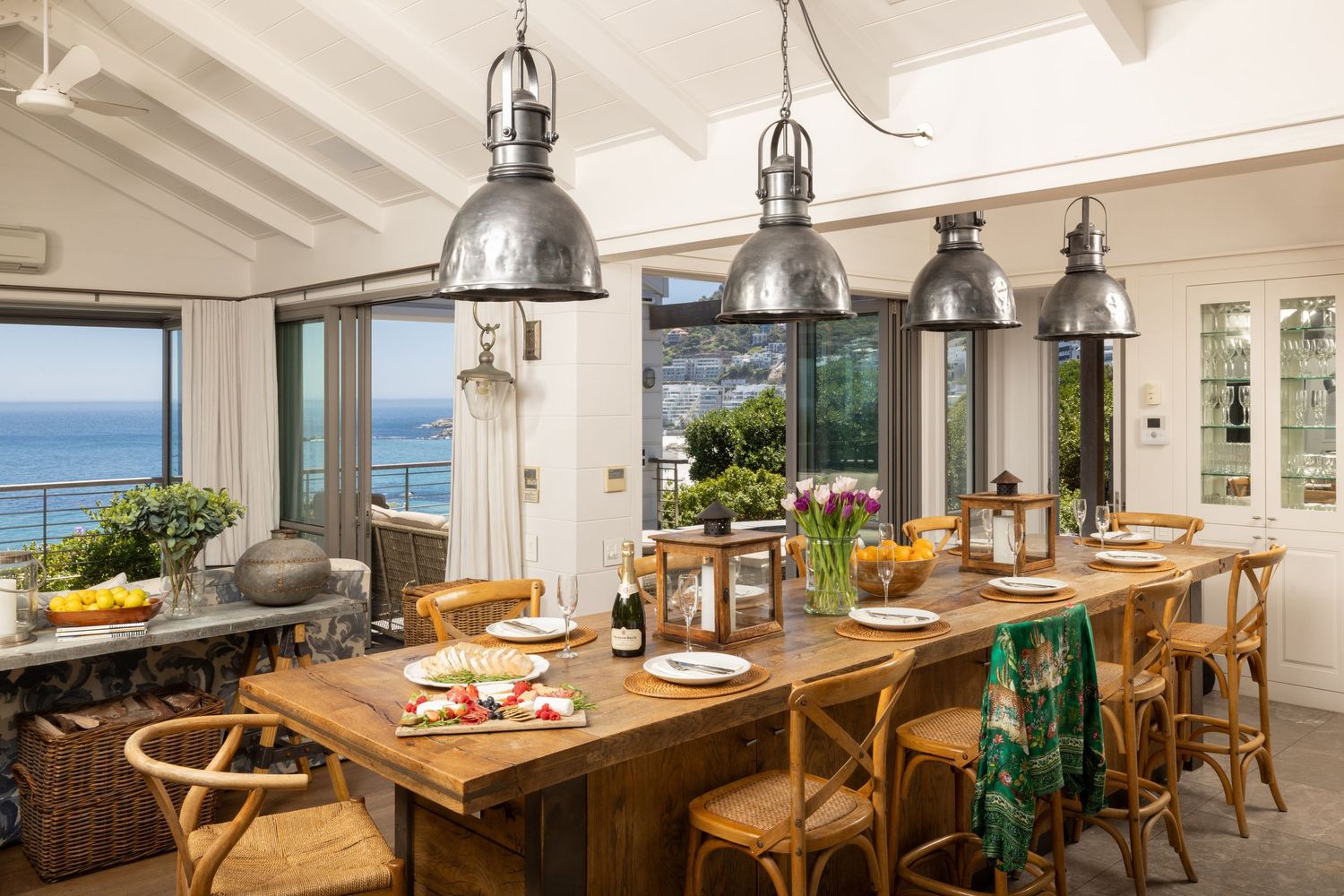 Coastal dining room with wooden table, rattan chairs, and pendant lighting overlooking ocean views through large windows.