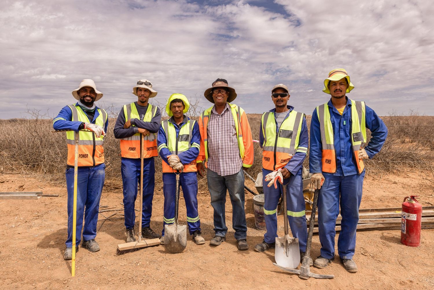 Construction crew members lined up at work site wearing blue uniforms and safety vests.
