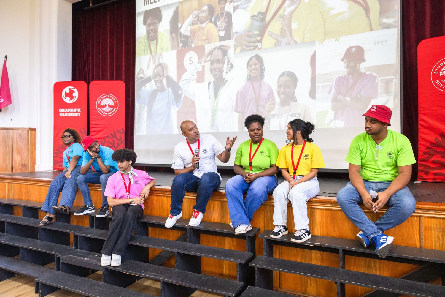 Participants in colorful shirts sit on benches during an indoor event with projector screens.