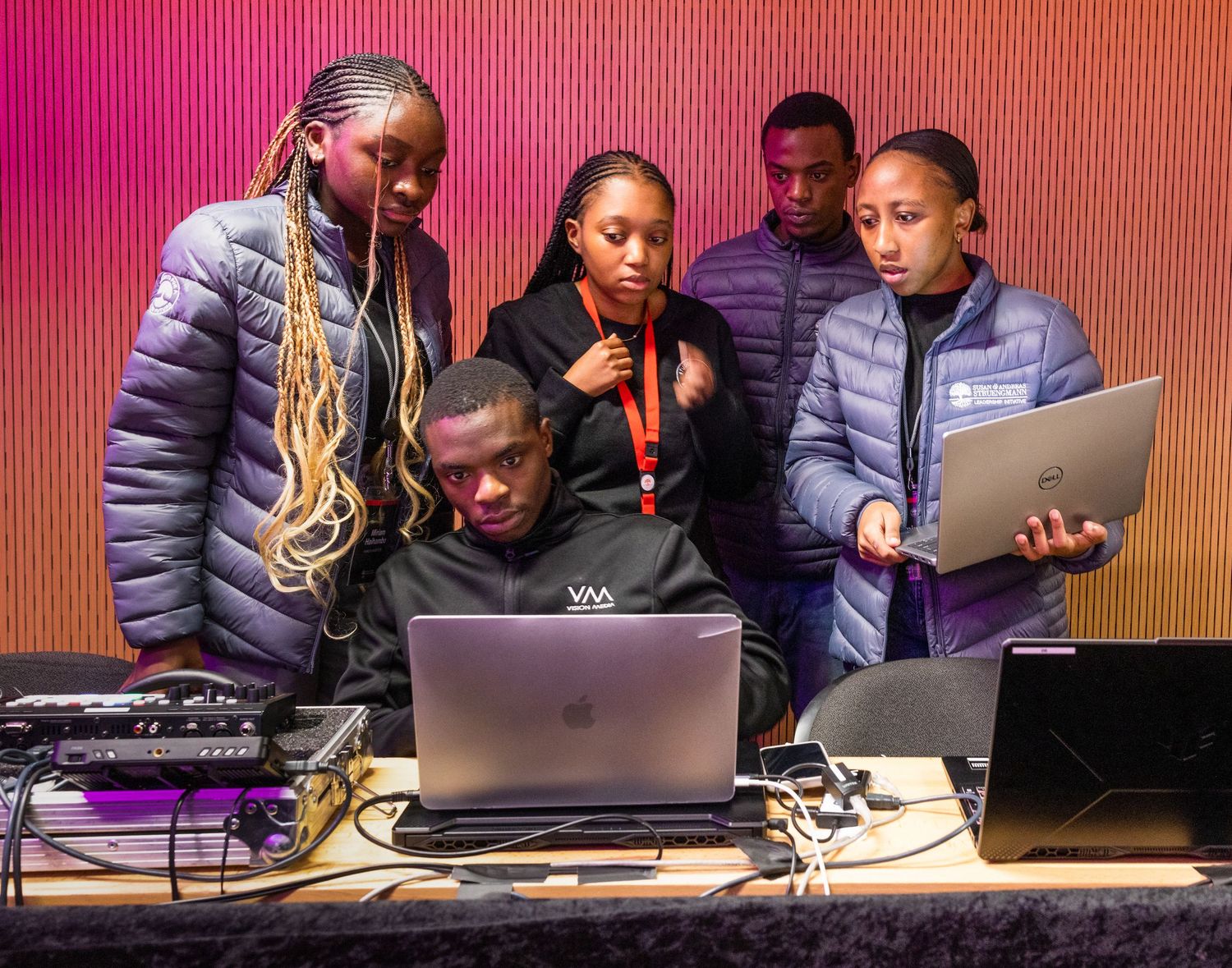Group gathered around laptops and audio equipment with colorful lighting in background.