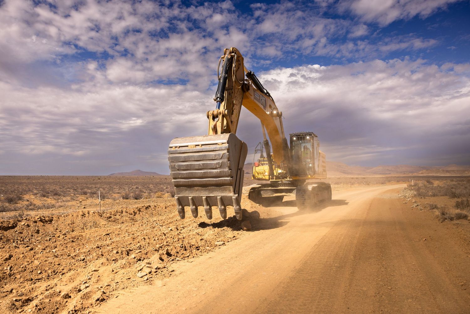 Heavy machinery kicks up dust while operating in desert terrain under cloudy skies.