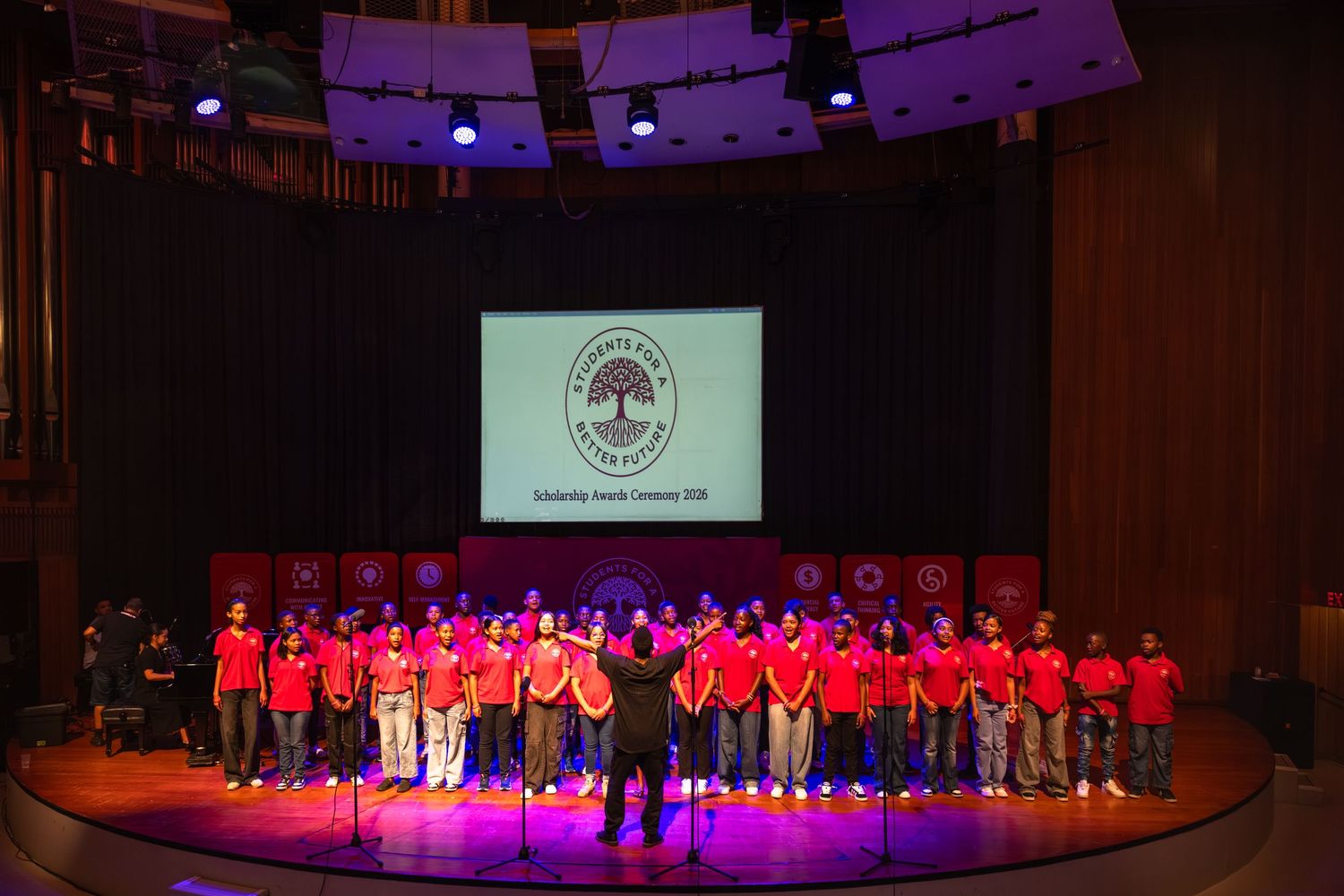 A group in red shirts performs on a dimly lit stage with a large screen displaying a logo in the background.
