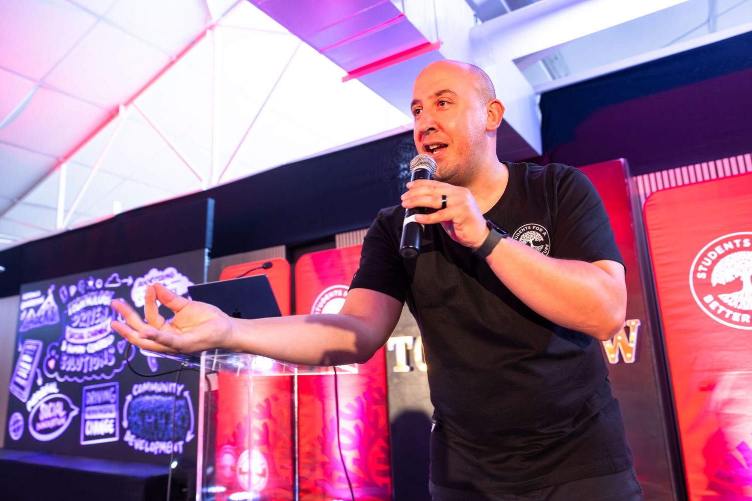 A speaker in a black polo shirt presents on stage with colorful lighting and branding visible in the background.