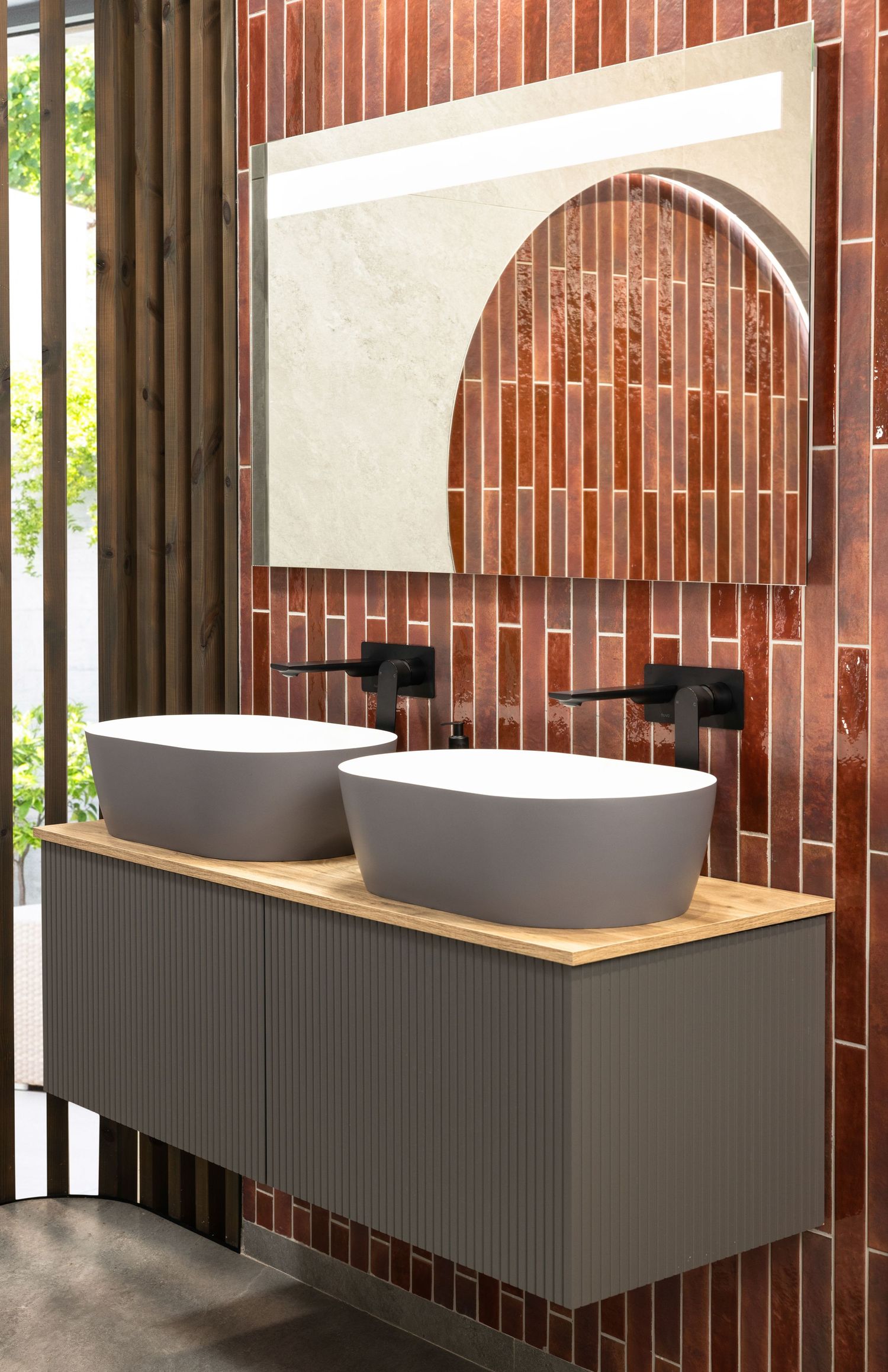 Modern bathroom vanity with two gray vessel sinks on wooden counter against terracotta tile wall with arched mirror.