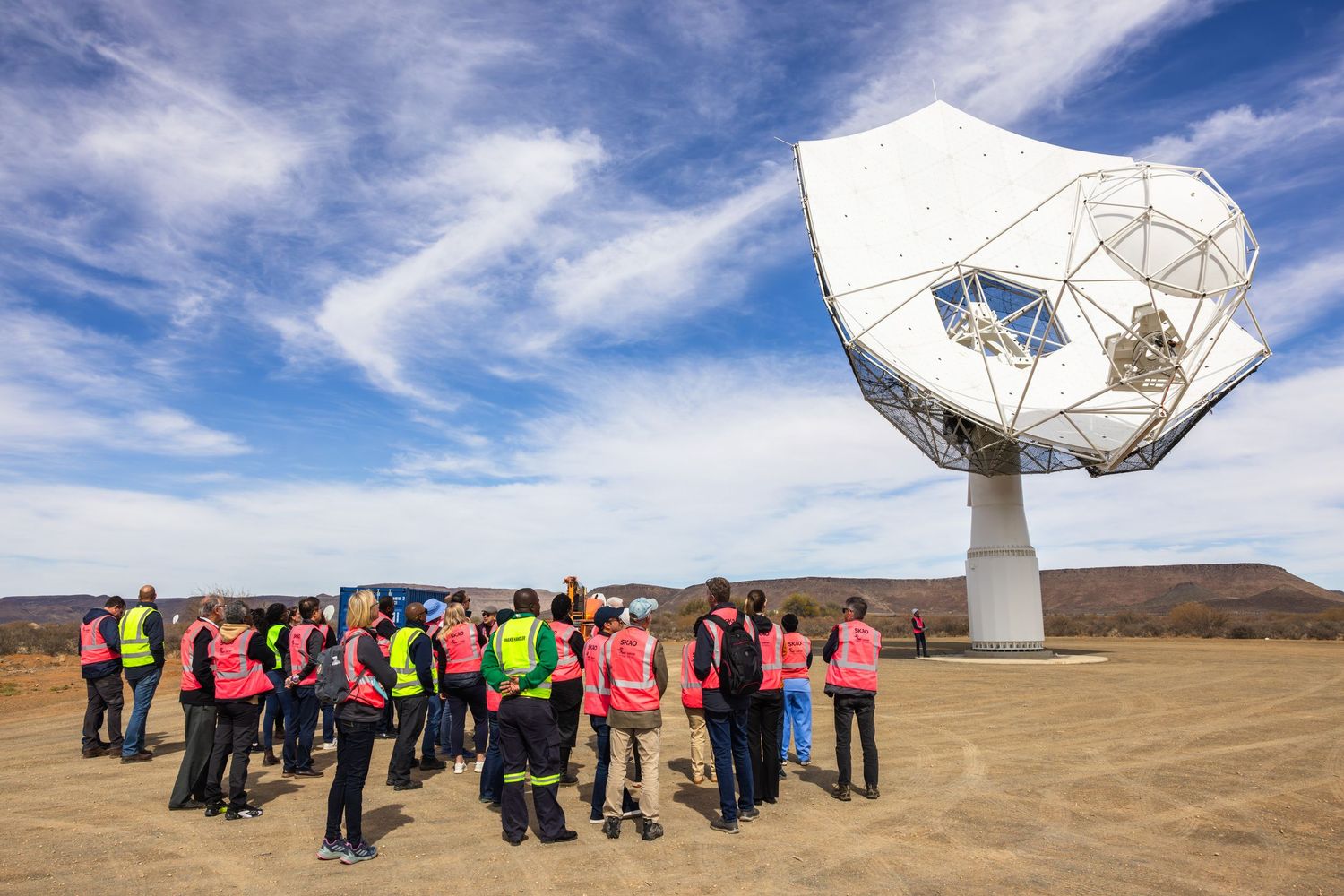 Large group of visitors in high-visibility vests gathered in front of a white radio telescope dish.