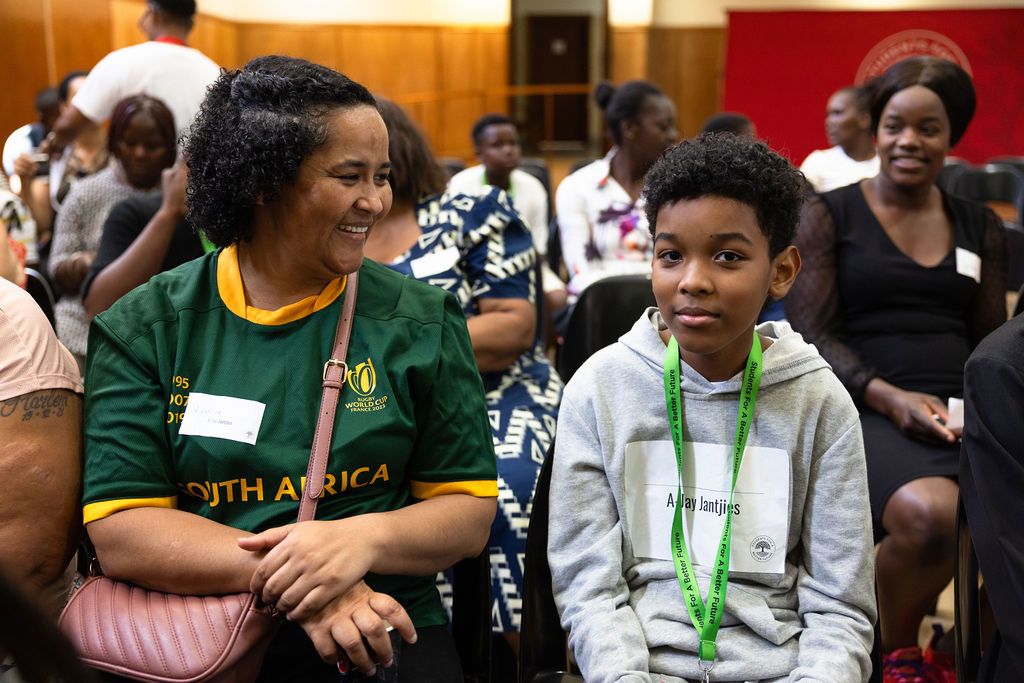 Two attendees at a community event smile while sitting together in an auditorium, one wearing a green jersey.
