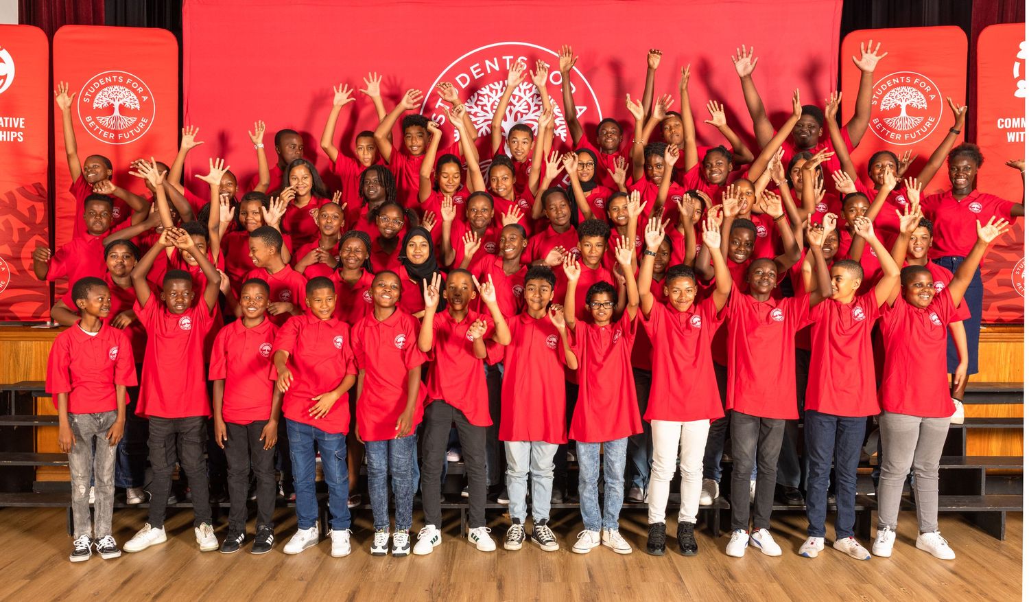 Large group wearing matching red shirts poses together against red background in school gymnasium.