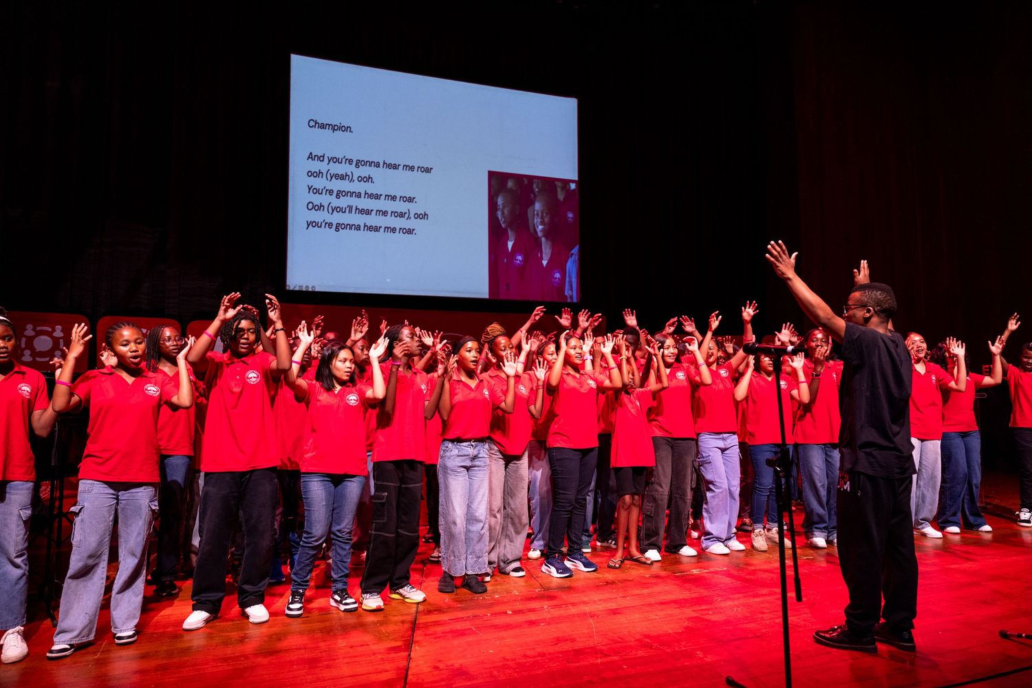 A group of performers in red shirts stand together on a brightly lit stage during a performance.