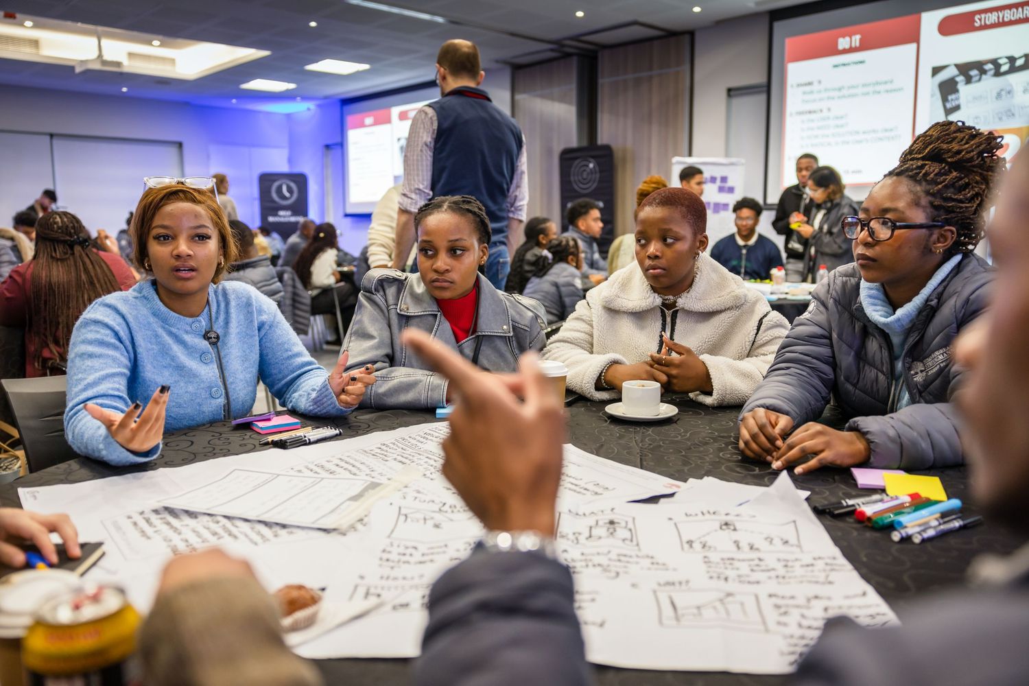 Students collaborate at a workshop table with papers and materials spread out during an educational session.