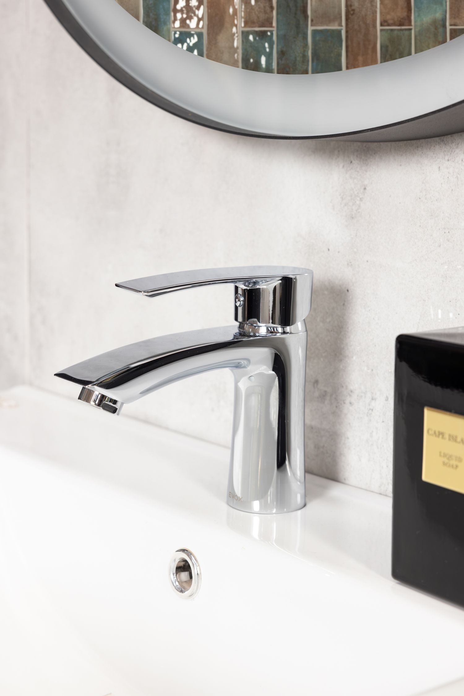 Modern chrome bathroom faucet mounted on white sink with round mirror above.