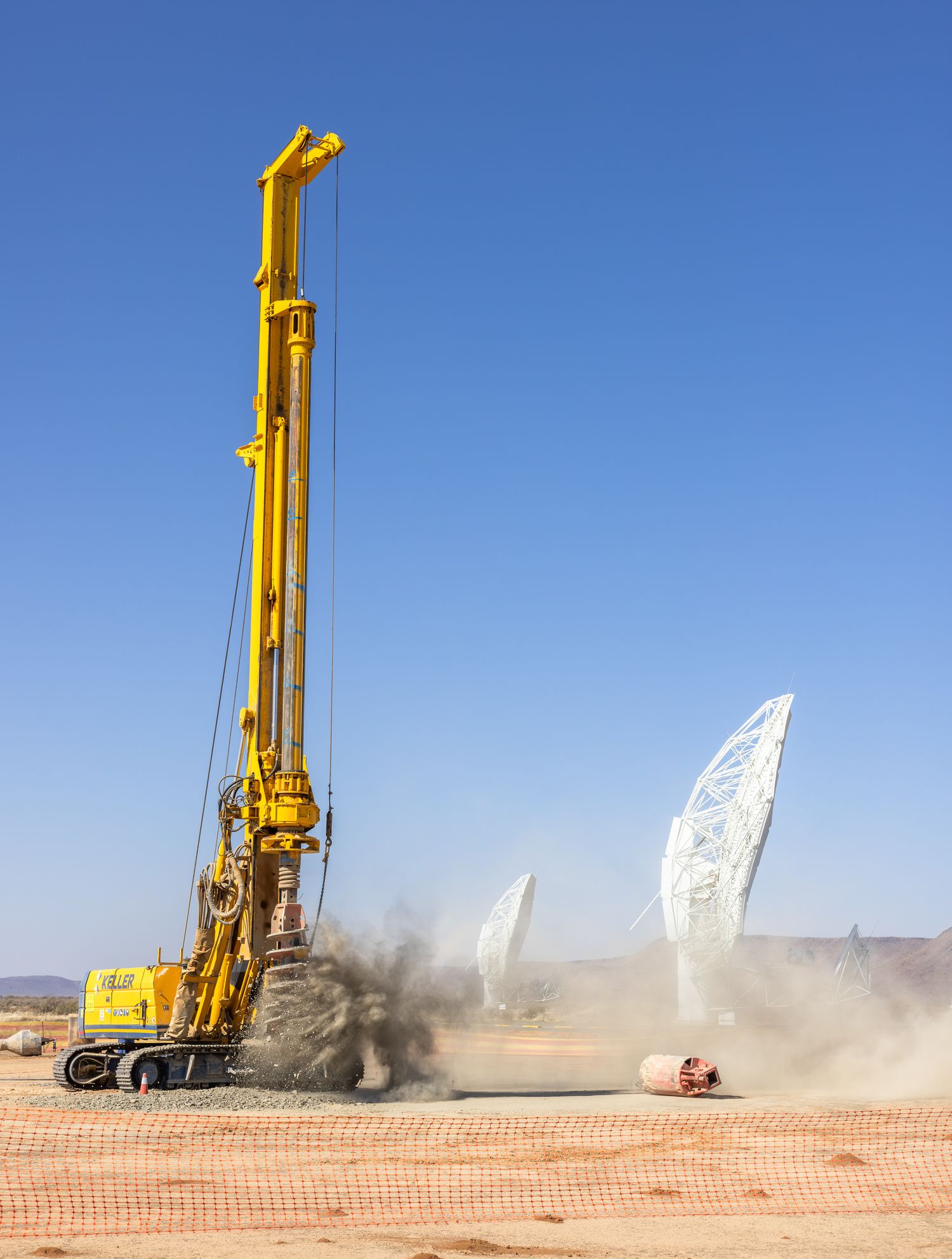 Yellow drilling rig operating in dusty desert conditions near white structures.