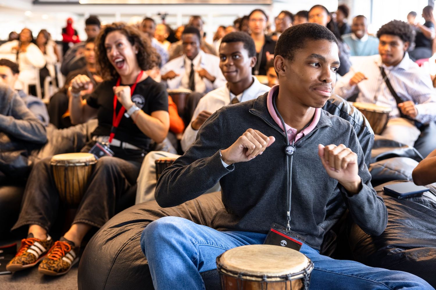 A diverse crowd participates in a drumming circle workshop with handheld percussion instruments.