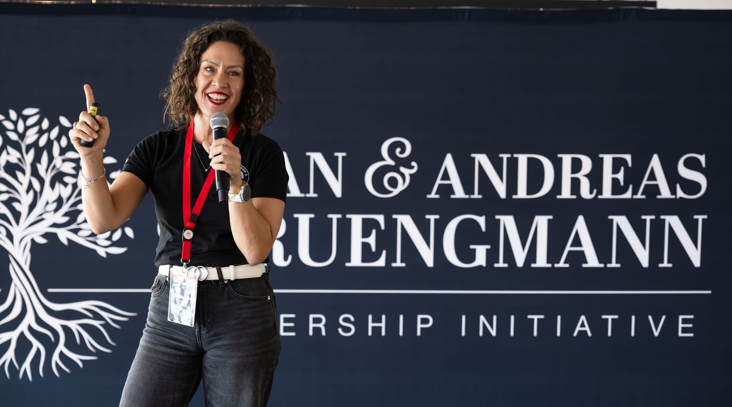 Speaker presenting on stage with microphone in front of a black backdrop with white text and tree logo.