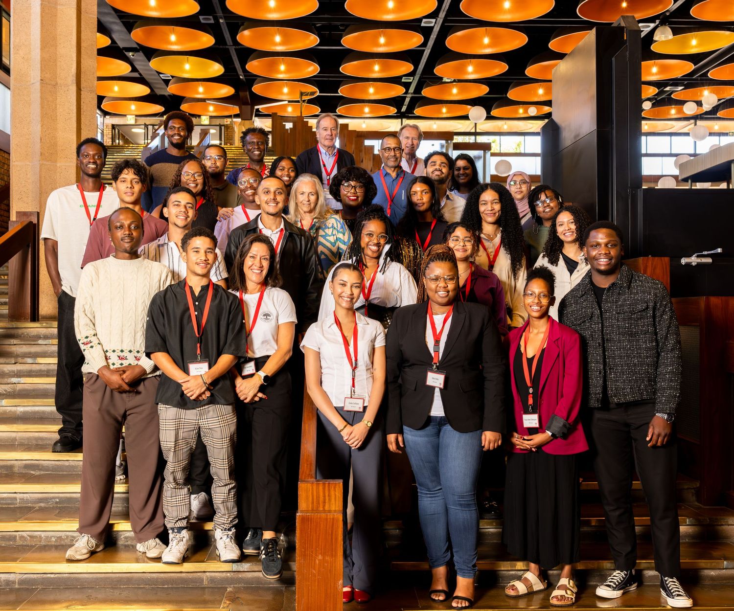 A diverse group of people pose together on stairs with orange circular lighting fixtures overhead.
