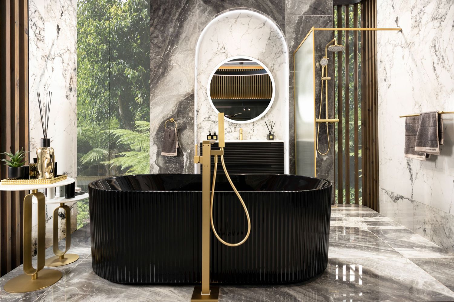 Modern black freestanding bathtub with brass fixtures in a luxurious marble bathroom with arched doorway and natural light.