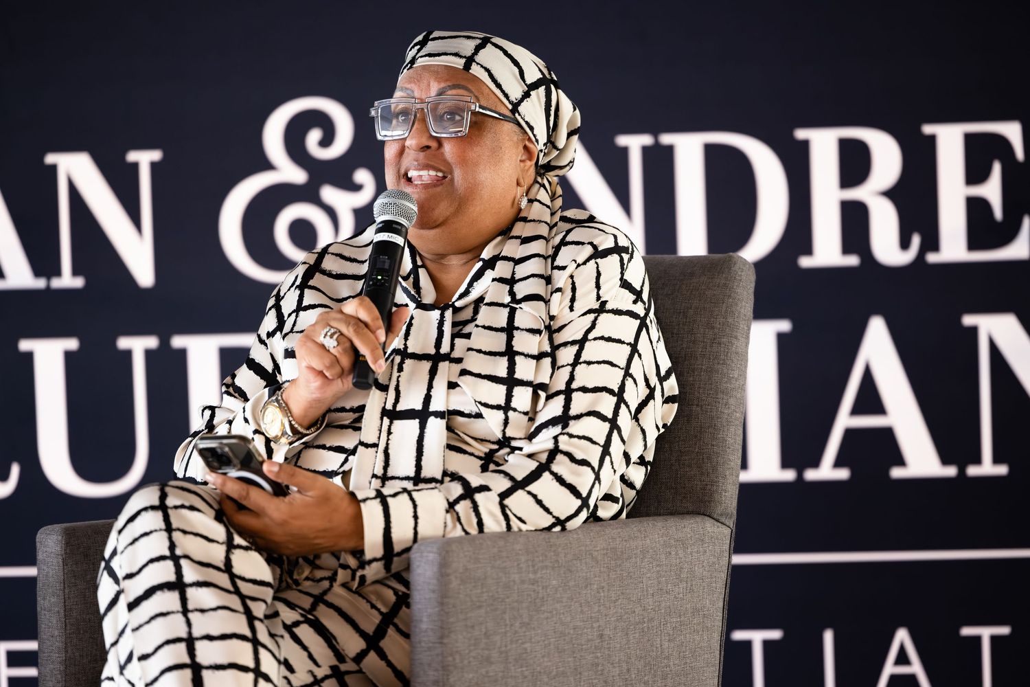Speaker in a geometric patterned outfit sits in a grey chair against a dark backdrop with text.
