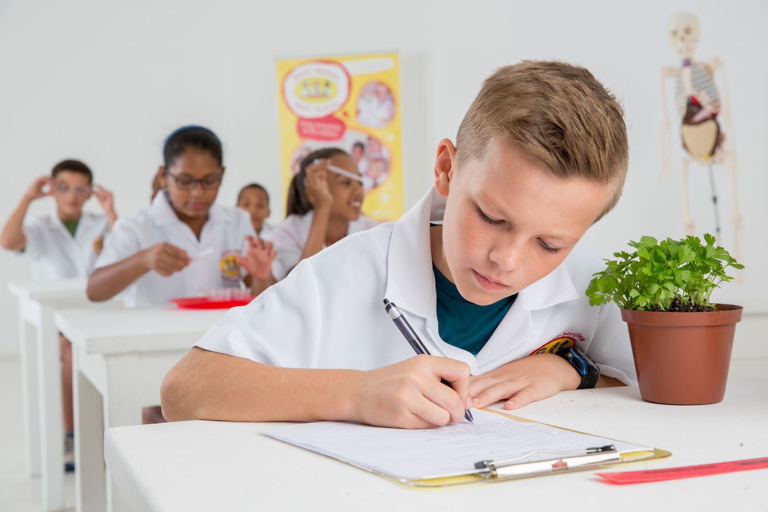 Student in white lab coat takes notes during science class while classmates work in background.