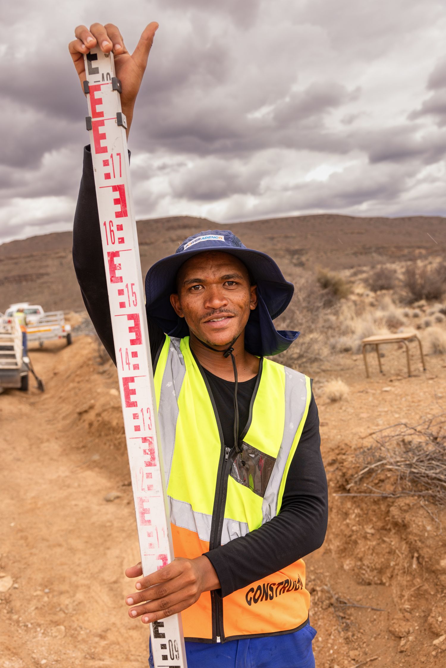 Construction worker holds measuring staff in desert terrain wearing safety gear.