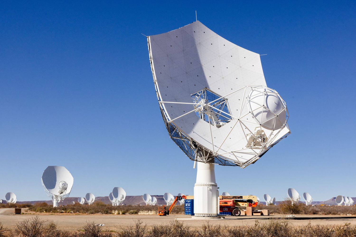 Large white radio telescope dish against blue sky with other satellite dishes in background.