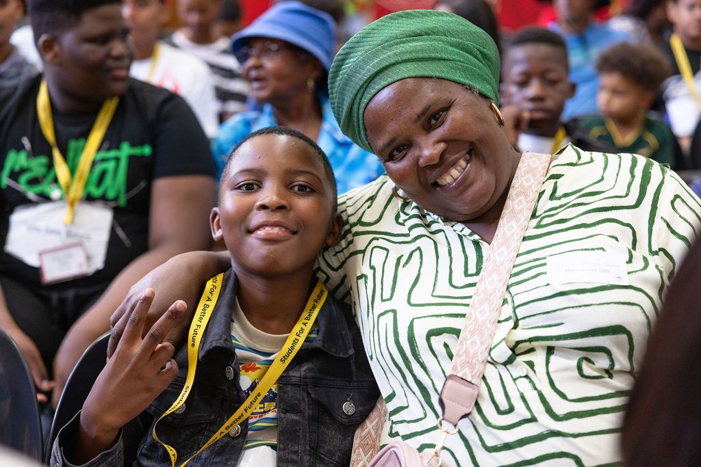 A diverse crowd of people wearing colorful clothing and headscarves gather together smiling and celebrating.