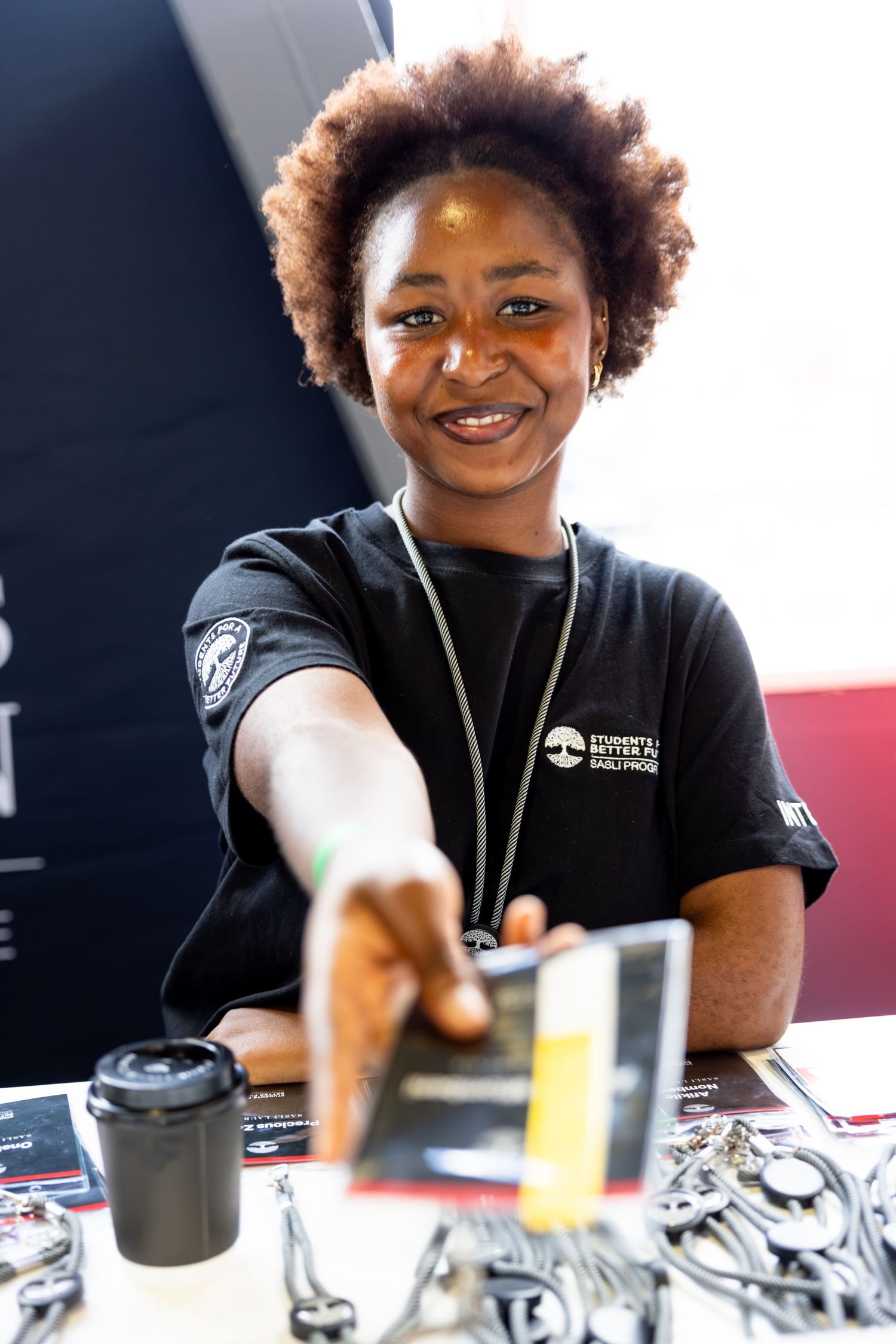 Staff member in black uniform shirt serves beverages at an event counter with drinks and promotional materials.