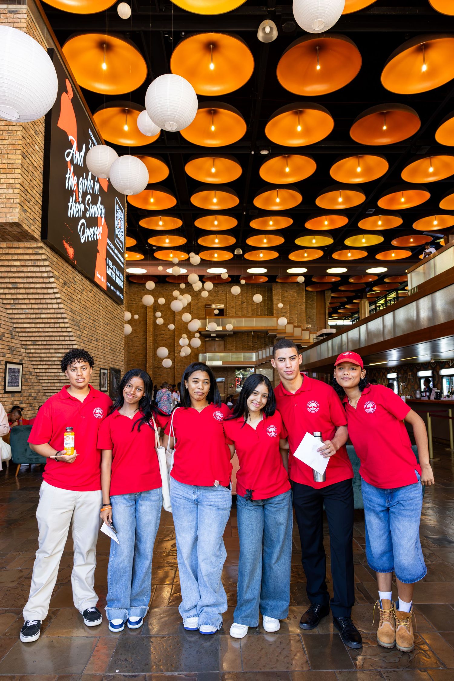 A group in matching red shirts and jeans poses together under orange circular ceiling lights in a hallway.