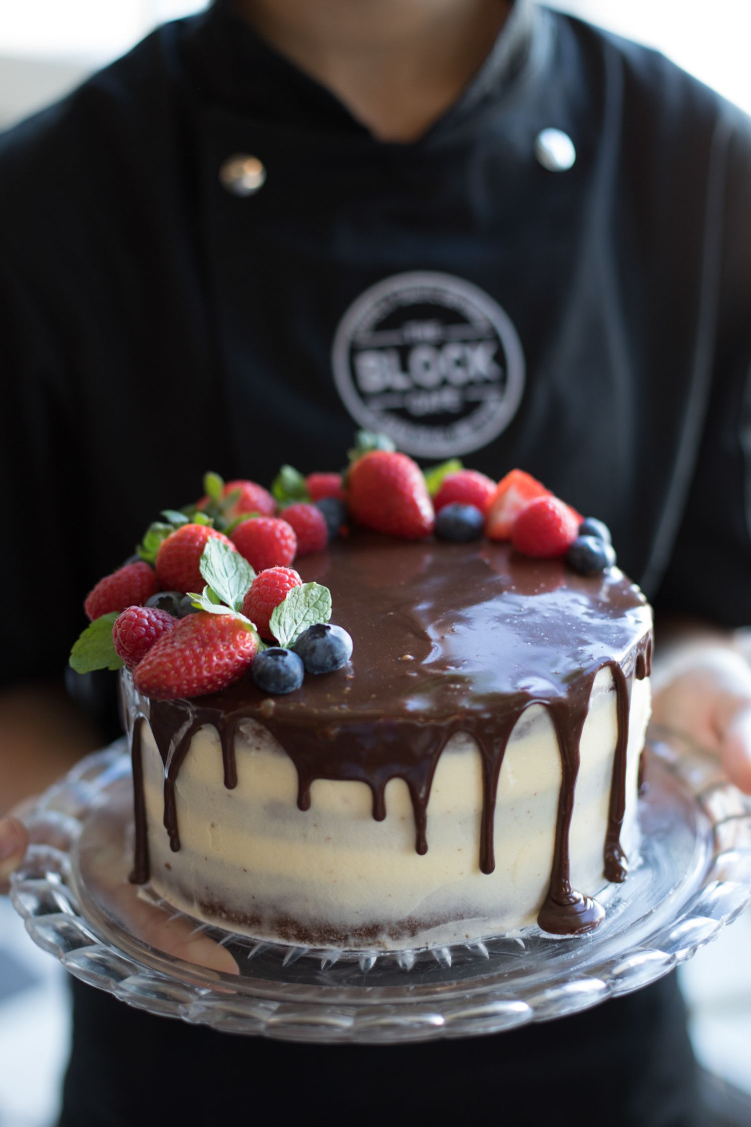 A cheesecake topped with chocolate ganache drip and fresh berries presented on a clear glass plate.