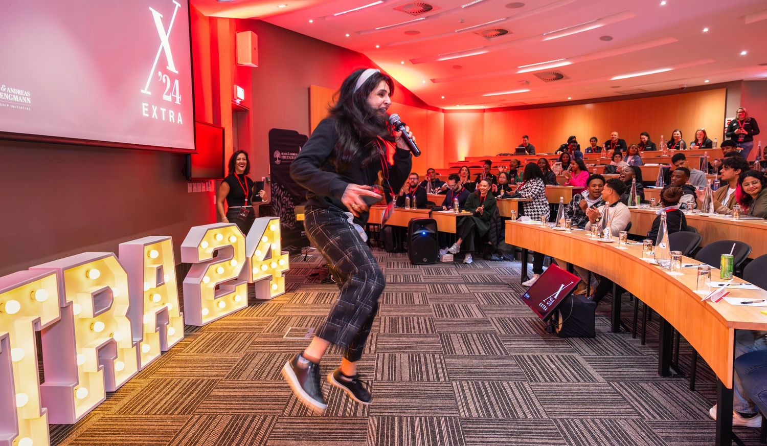 Energetic performer entertaining audience in conference room with red lighting and curved seating arrangement.