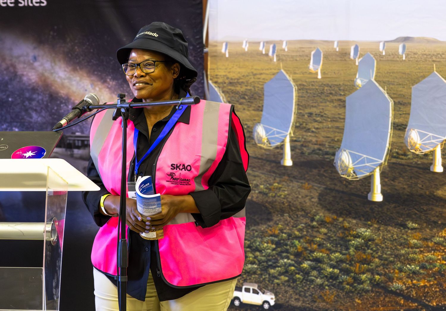 Safety-vested worker stands near astronomy equipment with radio telescope array in background.