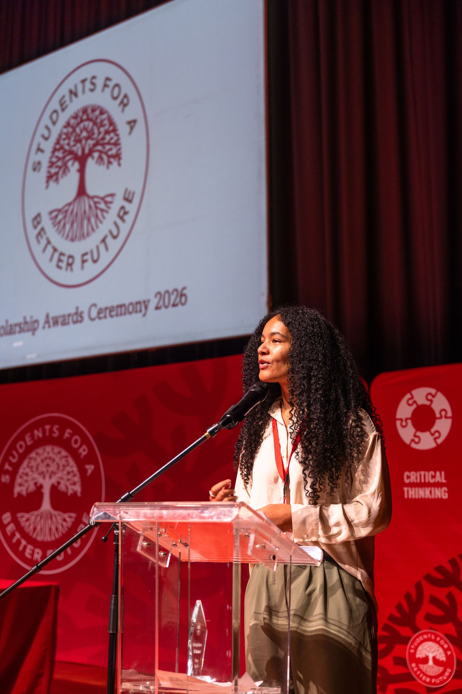Speaker at podium on stage with red backdrop and organizational tree logo visible.