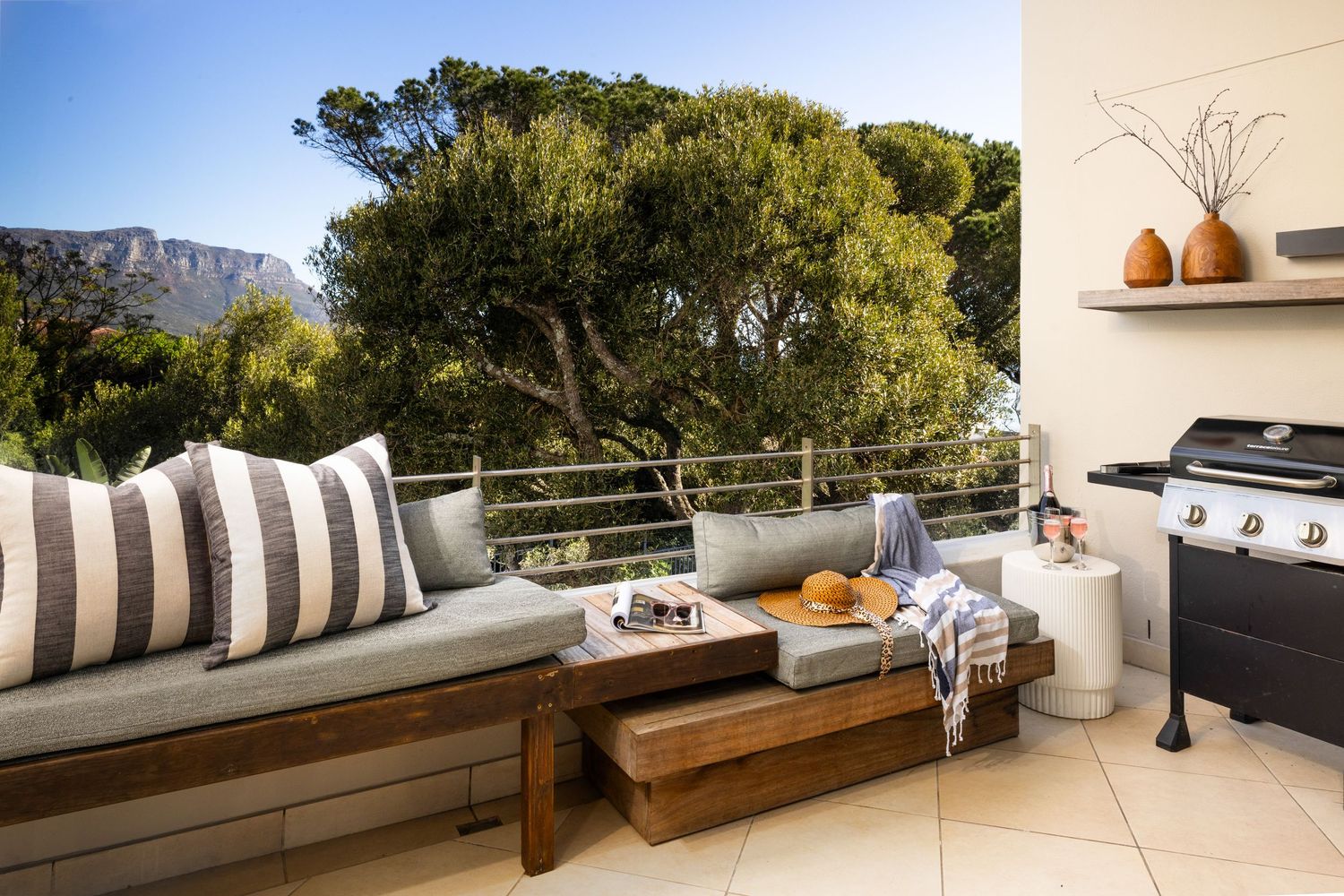 Modern outdoor patio with built-in bench seating, striped pillows, and a BBQ grill overlooking mountain scenery.