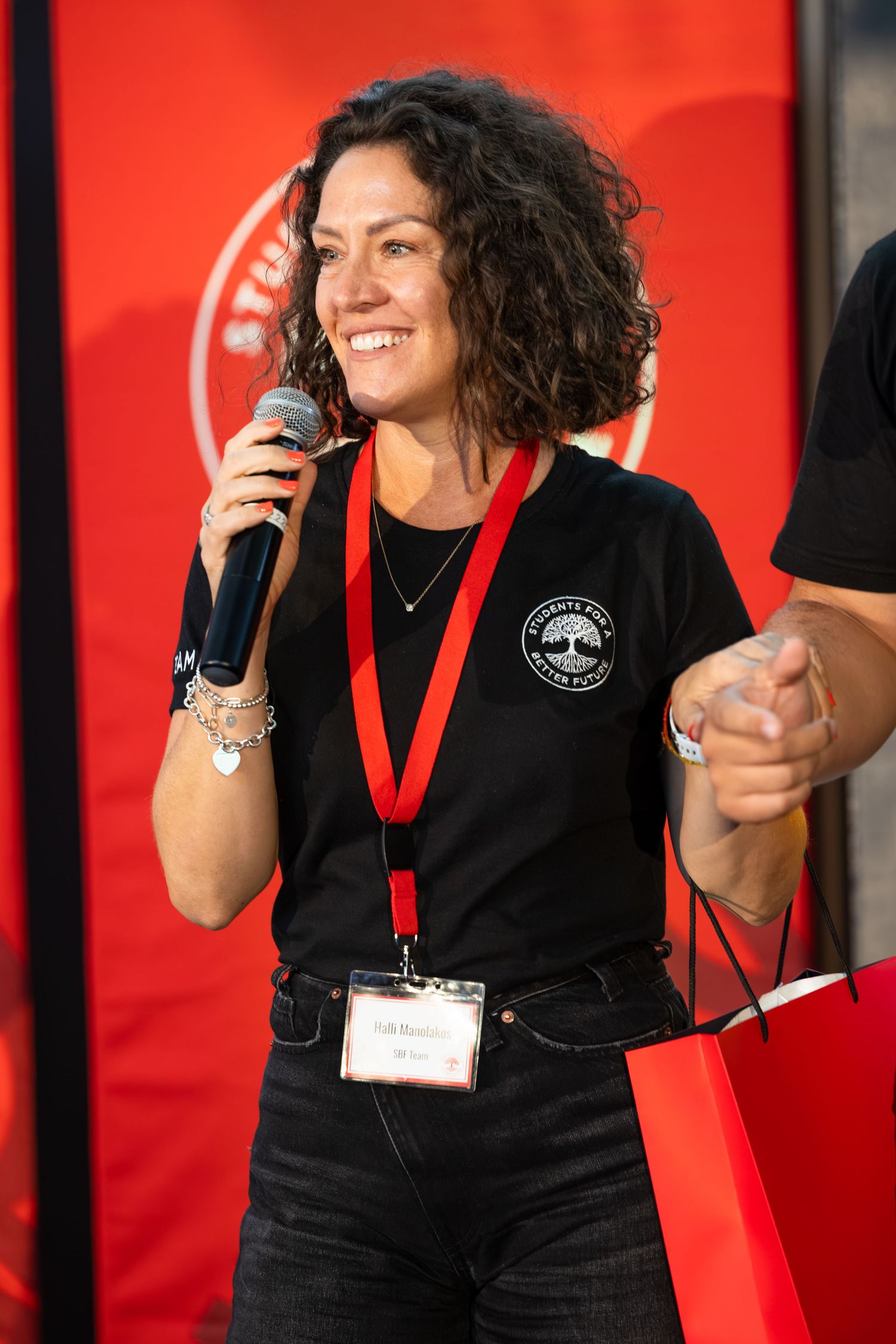A presenter wearing a black t-shirt with a lanyard speaks into a microphone against a red backdrop.
