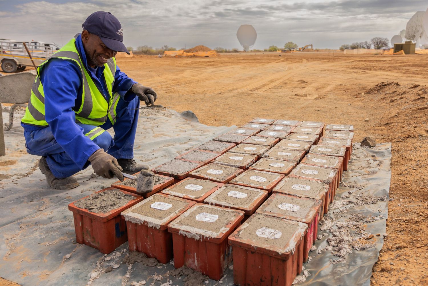 Worker laying bricks at construction site with satellite dishes visible in background.