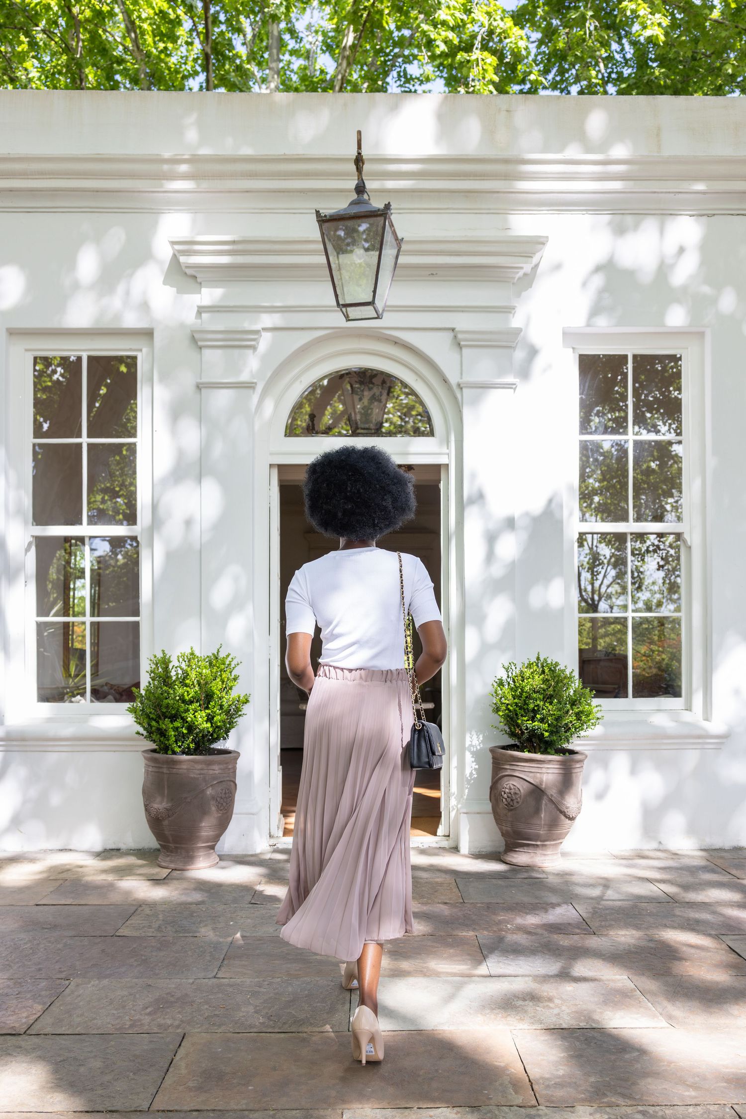 A person in a pleated pink skirt and white top approaches an elegant white colonial doorway with potted plants.