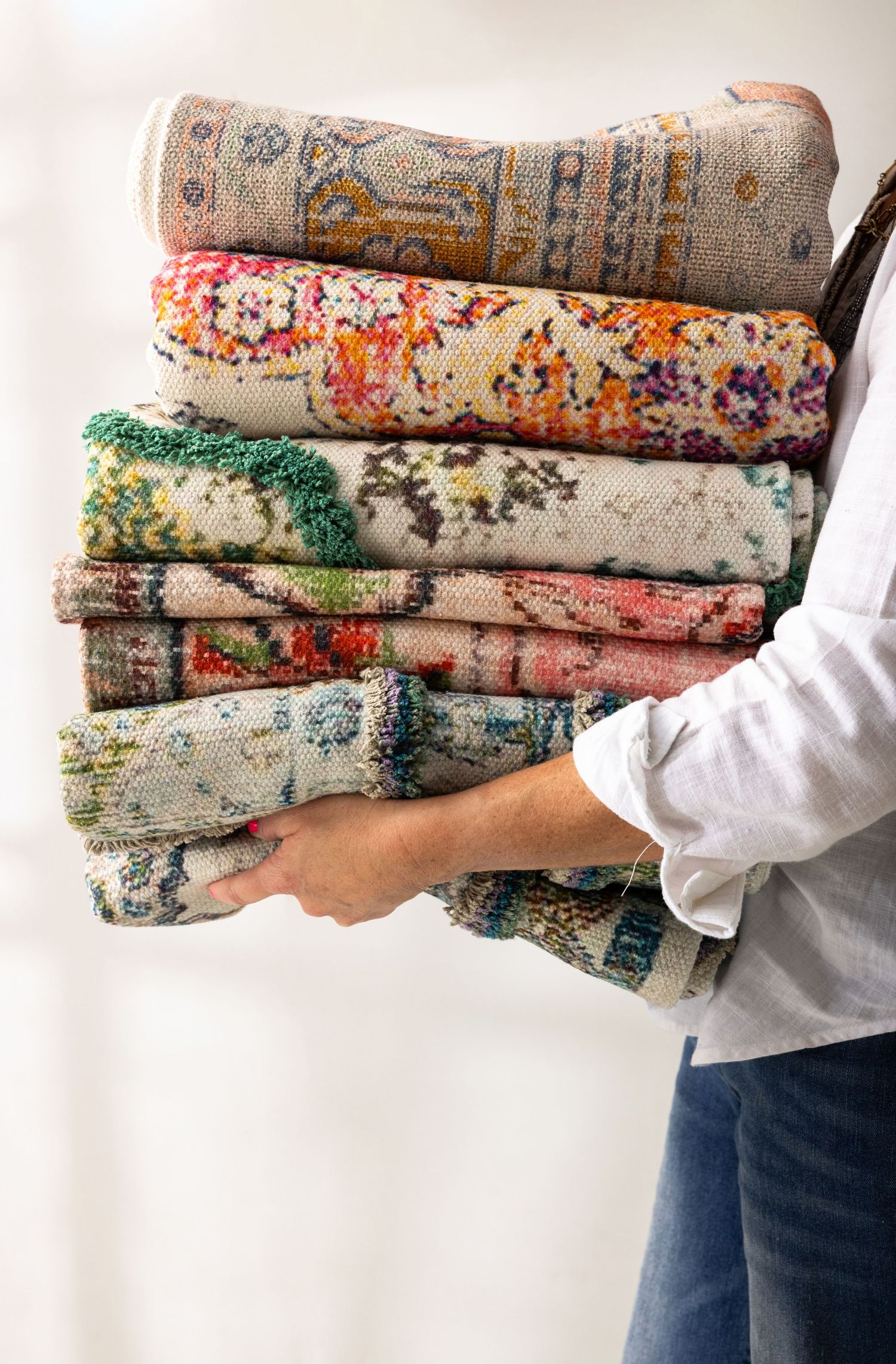 Stack of vintage floral rugs in muted colors being held by someone wearing a white sleeve and jeans.