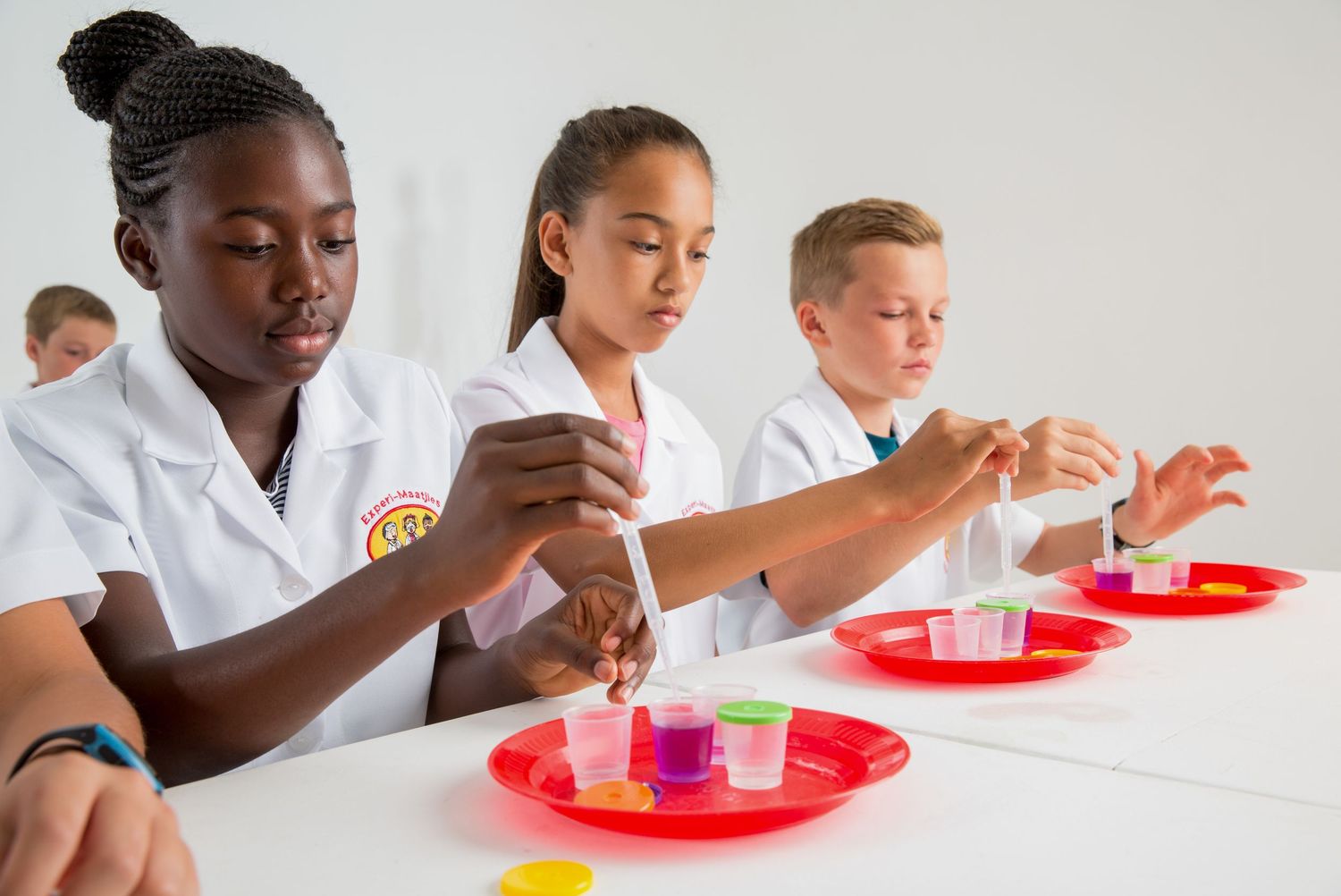 Students in white lab coats conduct chemistry experiments with test tubes and red plates on a laboratory table.