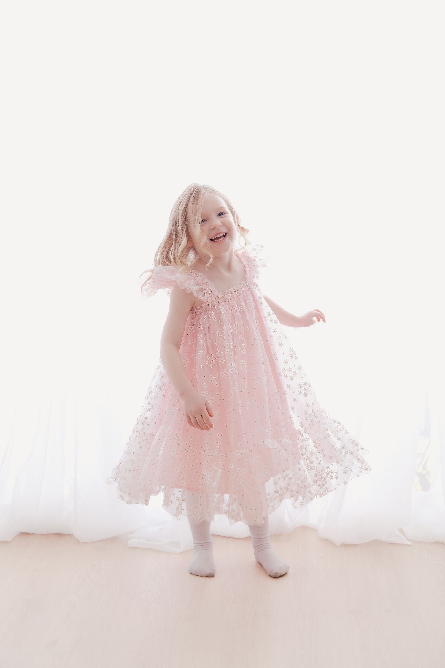 A joyful young girl twirls in a pink lace dress in a bright white studio.