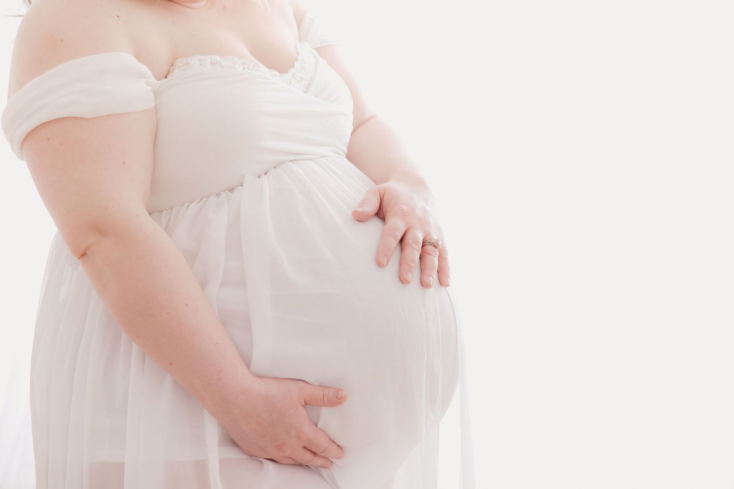 Pregnant woman in white dress cradles her belly against a bright studio backdrop.